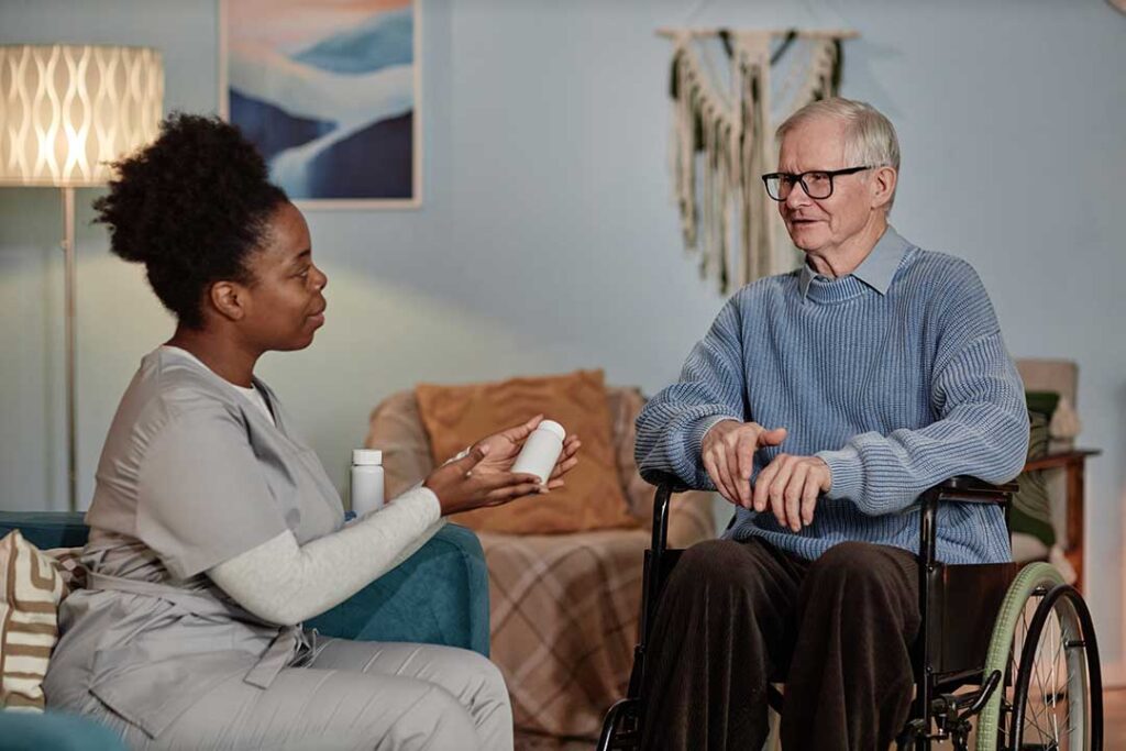 An older adult receives medication support from a caregiver in a cheerful, home-like common room.